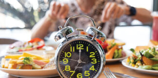 Selective Focus Of Alarm Clock With Young Man Eating A Healthy Food As Intermittent Fasting, Time Restricted Eating Diet Breakfast Image