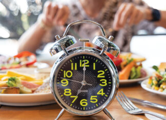 Selective Focus Of Alarm Clock With Young Man Eating A Healthy Food As Intermittent Fasting, Time Restricted Eating Diet Breakfast Image