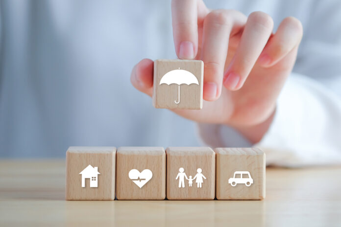 Insurance Concept. Protection Against A Possible Eventuality. Hand Holding Umbrella Icon And House, Car, Family And Health Icon On Wooden Block For Assurance Life Concept.
