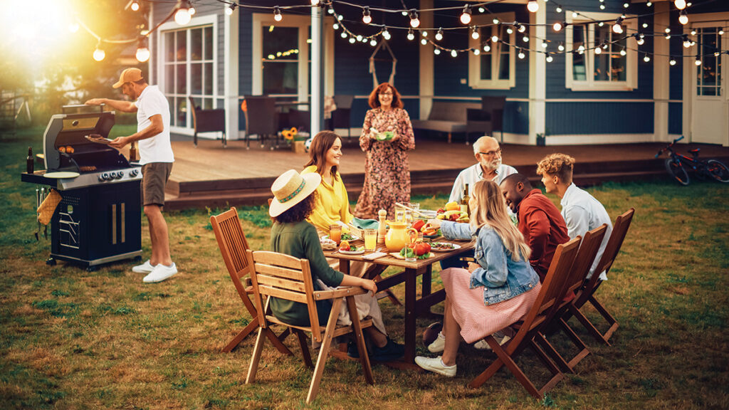 Family And Multiethnic Diverse Friends Gathering Together At A Garden Table. People Cooking Meat On A Fire Grill, Preparing Tasty Salads For A Big Family Celebration With Relatives.