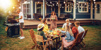 Family And Multiethnic Diverse Friends Gathering Together At A Garden Table. People Cooking Meat On A Fire Grill, Preparing Tasty Salads For A Big Family Celebration With Relatives.