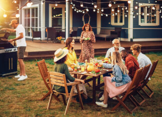 Family And Multiethnic Diverse Friends Gathering Together At A Garden Table. People Cooking Meat On A Fire Grill, Preparing Tasty Salads For A Big Family Celebration With Relatives.