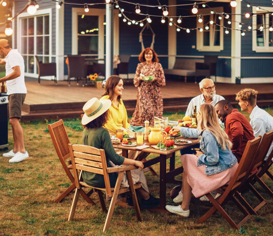 Family And Multiethnic Diverse Friends Gathering Together At A Garden Table. People Cooking Meat On A Fire Grill, Preparing Tasty Salads For A Big Family Celebration With Relatives.