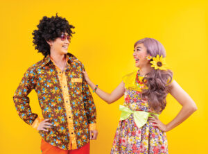 Asian Hippie Couple Dress In 80s Vintage Fashion With Colorful Retro Clothing While Dancing Together Isolated On Yellow Background For Fancy Outfit Party And Pop Culture